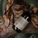 Oway Curly Hair Bath 240ml bottle resting diagonally on bark among dried leaves